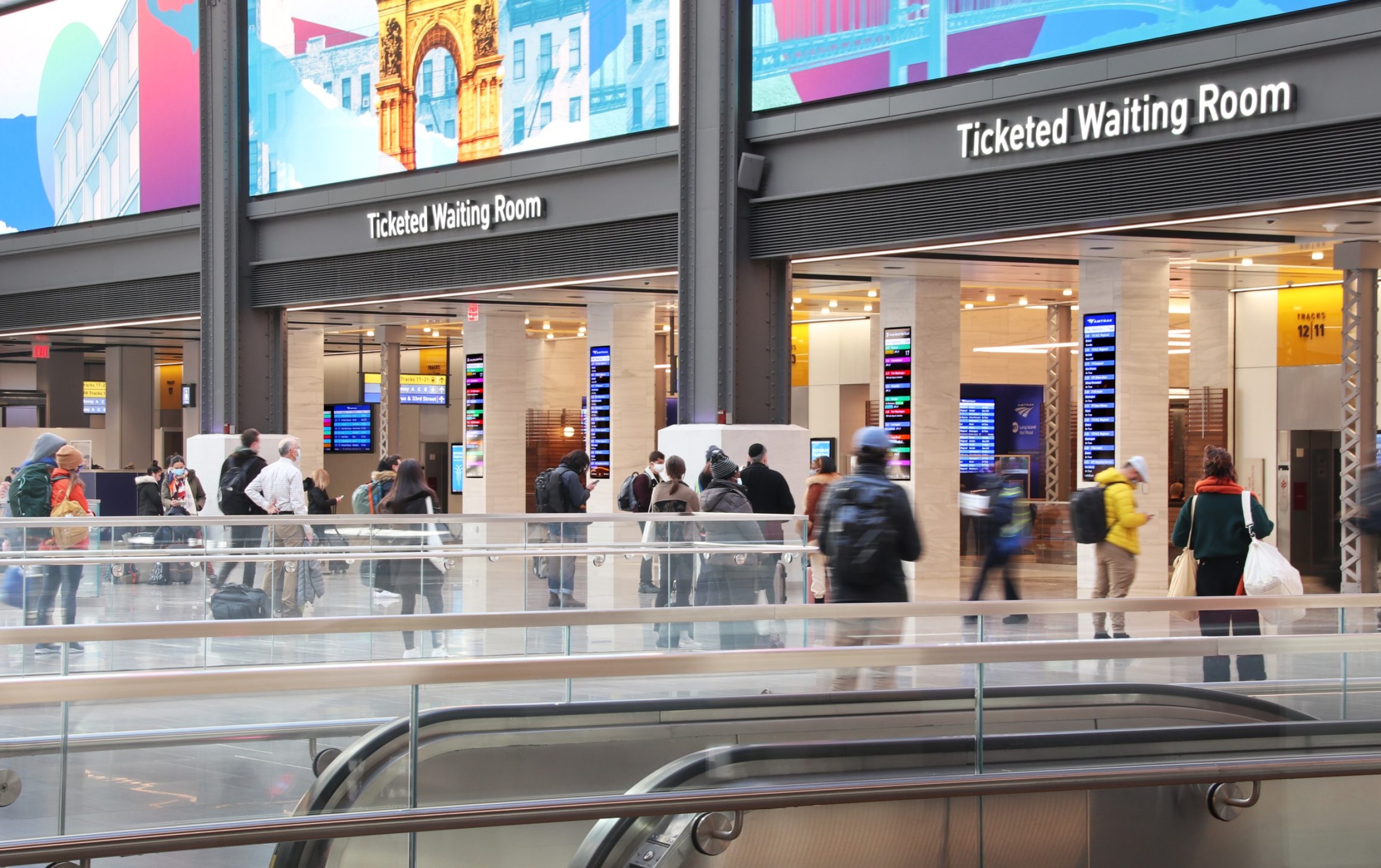 From just behind two escalators, we see another view of the Moynihain train station, as travellers walk through an entrance into what is displayed in illuminated letters above the entrance, as a ‘ticketed waiting room.’ Above the letters are colorful graphics of destinations, such as the Paris Arc du Triomphe. Travelers walk through tall beige pillars that hold the structure, gazing towards colorful guidelines on a screen in the pillars.