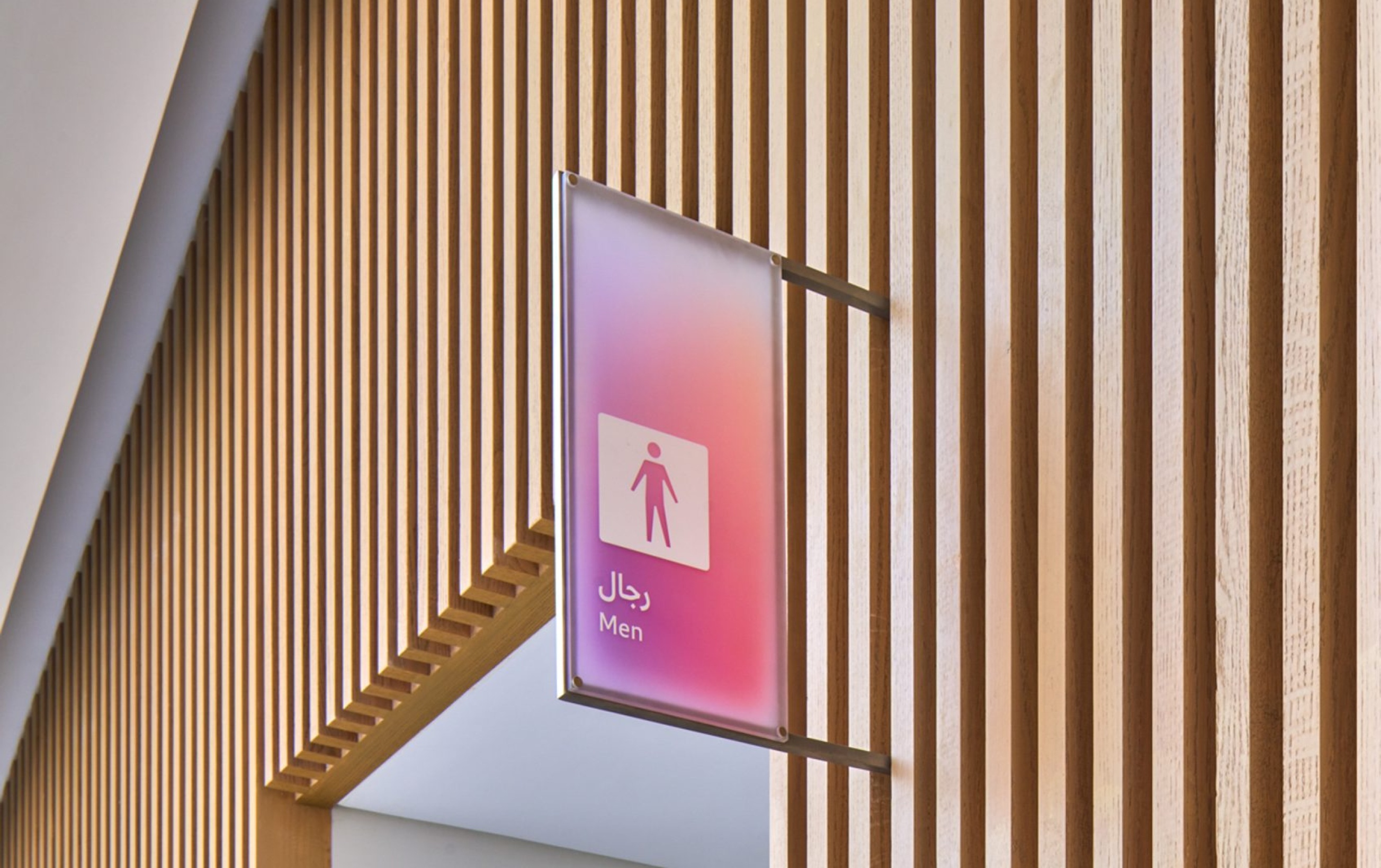 A photograph taken from a worm's-eye view of a wooden slatted wall with an opening that serves as a door. A pink and purple colored wayfinding sign is mounted between two slats to identify the male toilet. The sign is made of a milky, half-transparent material and is mounted on a brushed aluminum frame.