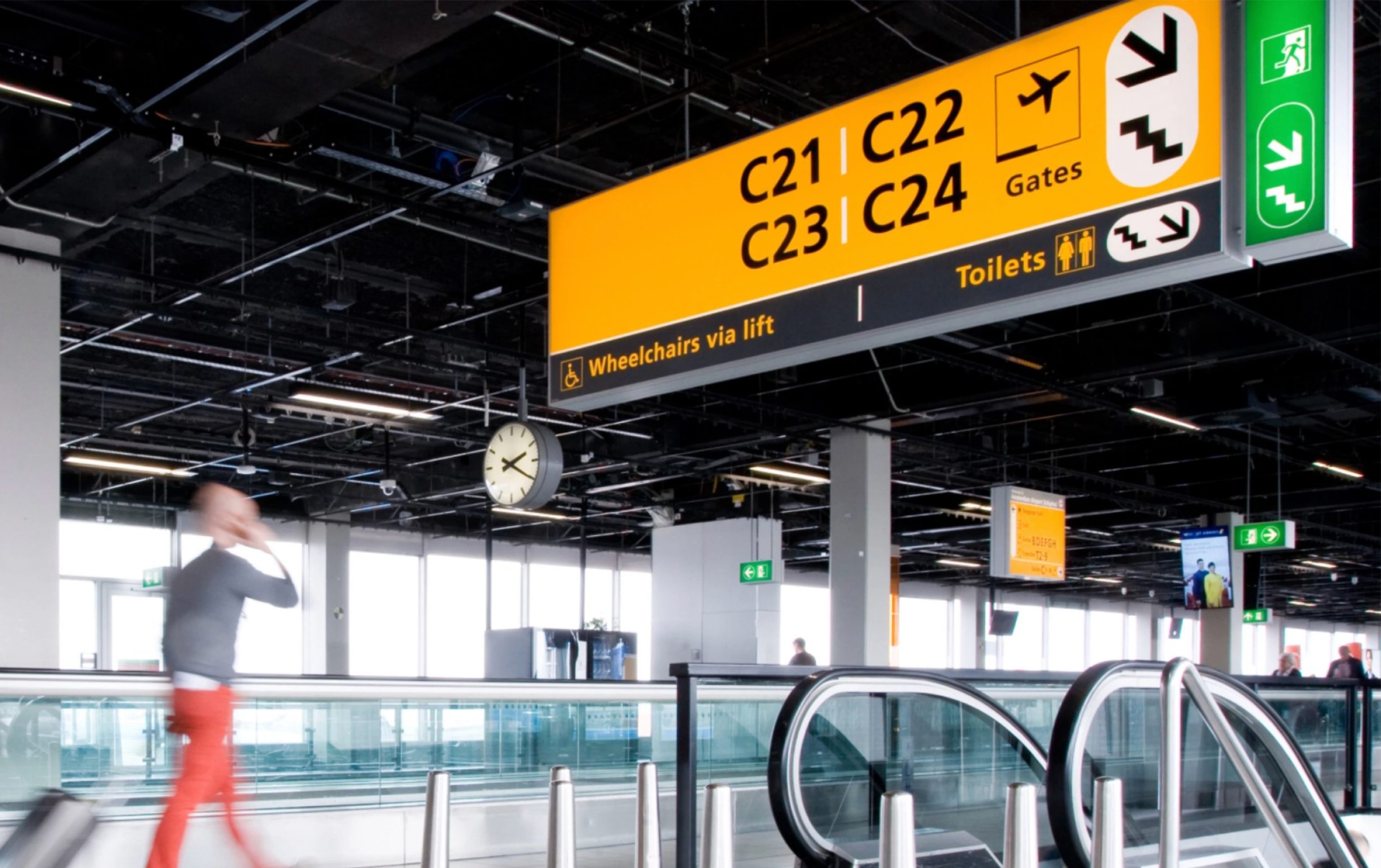 A wide-angle shot of a blurry man talking on the phone, walking towards a yellow sign displaying gates, directions, a wheelchair path, and the exits in Schiphol airport.