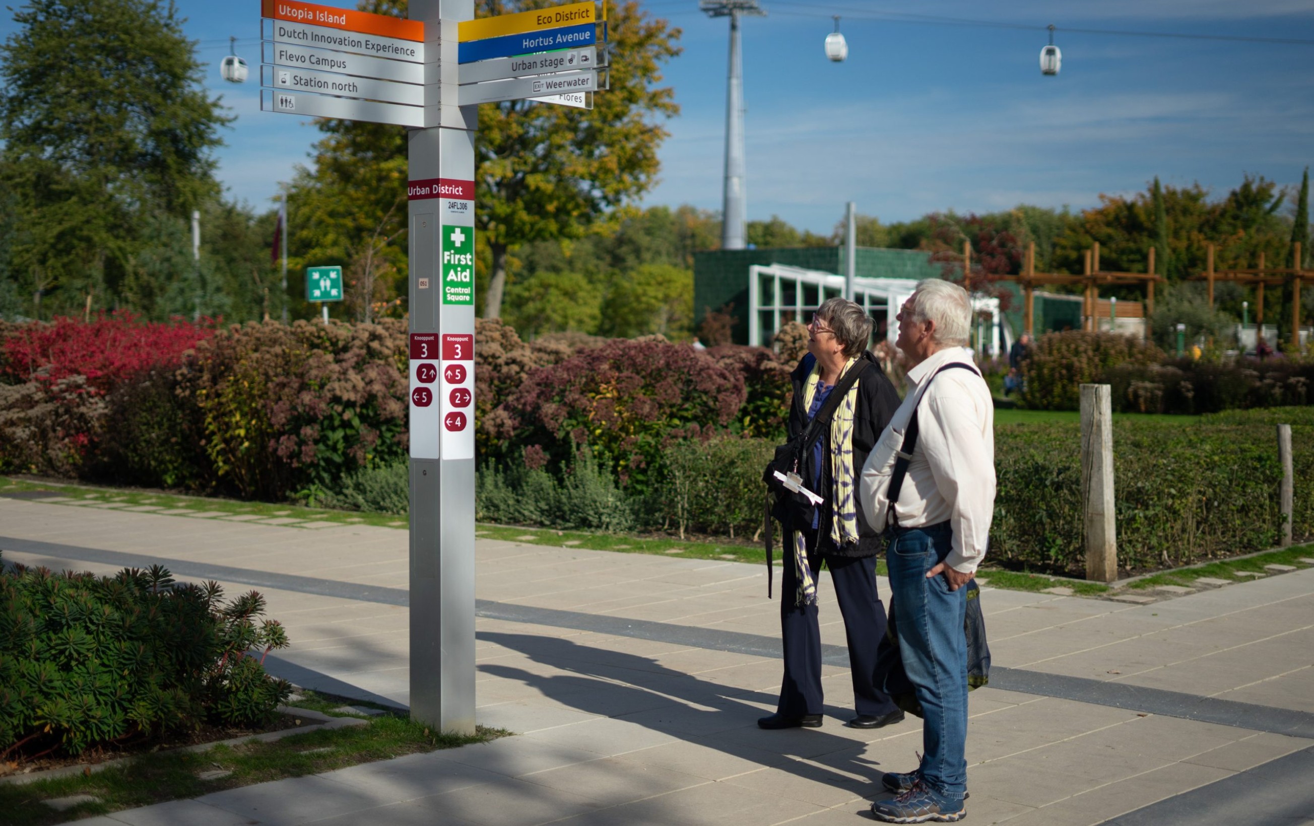 A photograph taken at eye-view of an elderly male and female couple looking up at a finger pointer designed by Mijksenaar at the Floriade Expo. It is a sunny day with, in the background, blue skies, a few clouds, and a cable cart. Along the horizon, behind the couple, there are trees and bushes in many different colors and varying sizes and a green and a small, green and white building, one story high with multiple window panes.
