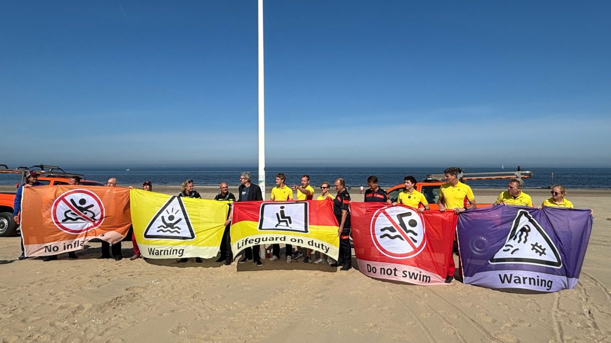 16 mensen staan op een rij op het strand en houden samen vijf gekleurde vlaggen met teksten en pictogrammen omhoog. Foto credit: Reddingsbrigade Nederland.